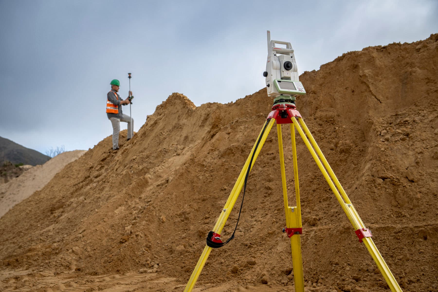 A Leica robotic total station on a tripod, with a surveyor taking a point on the stockpile behind it.